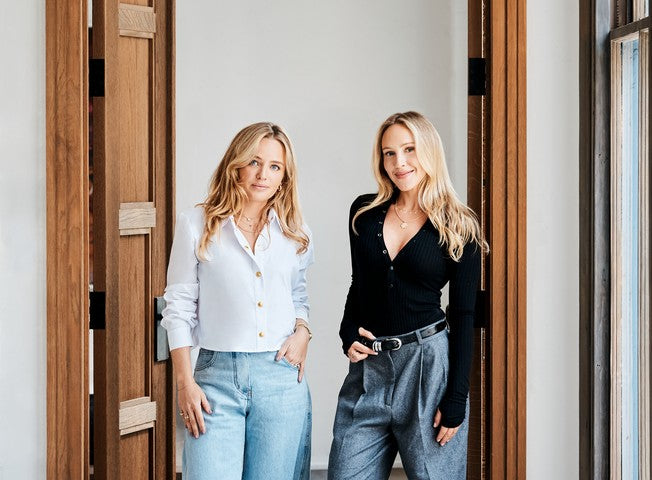 Two women standing in a doorway wearing casual outfits. The woman on the left wears a white button-down shirt and light blue jeans, while the woman on the right wears a black long-sleeve top and gray wide-leg jeans.