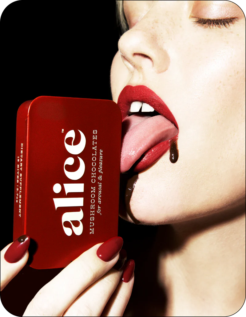 A close-up lifestyle shot of a woman with red lipstick and red nails holding a red tin of alice Mushroom Chocolates, with chocolate dripping from her mouth against a black background.