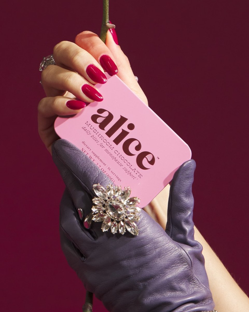 A hand in a lavender glove holding a pink Menopause Hero tin and a pink orchid on a magenta backdrop
