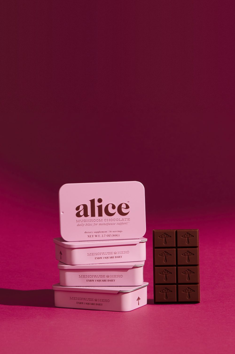 Three pink Menopause Hero tins stacked beside a bar of alice dark chocolate on a magenta studio backdrop