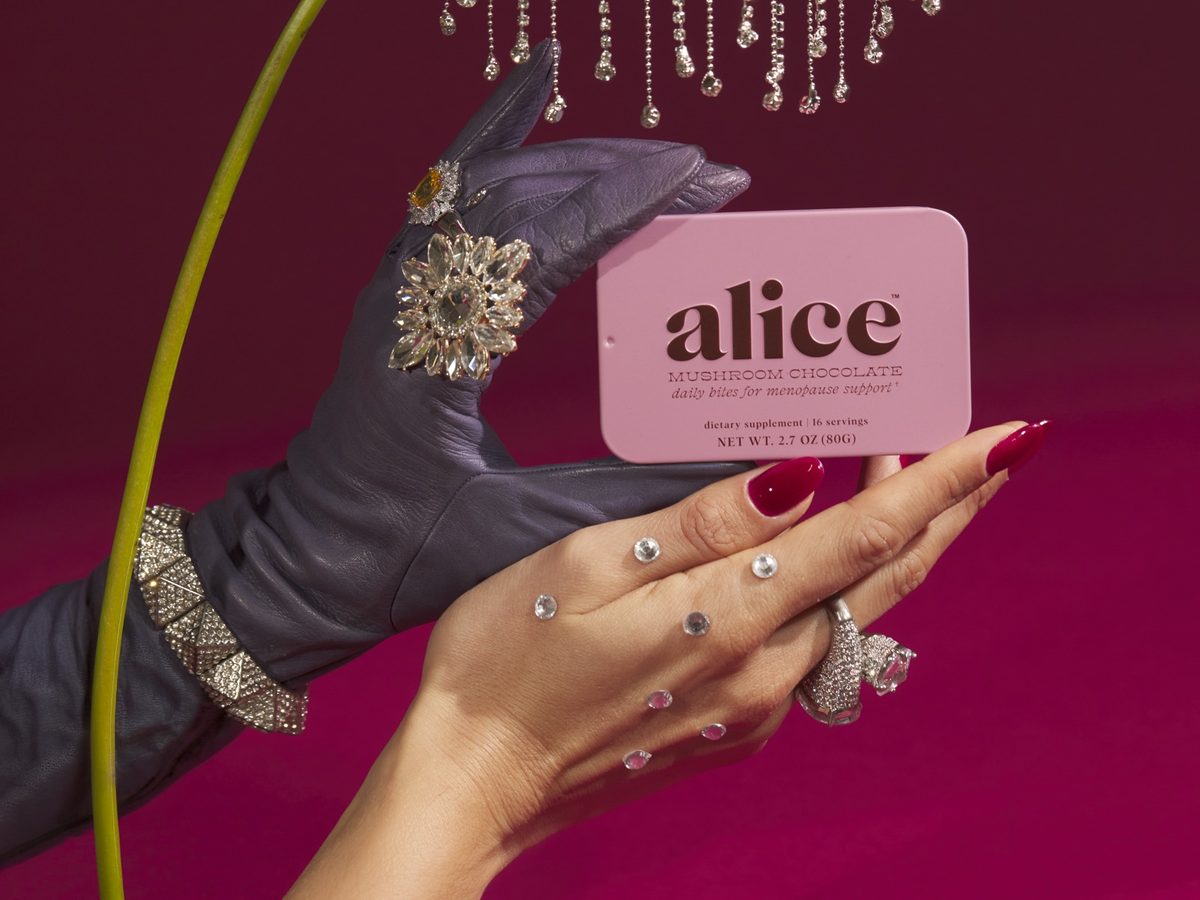 A gloved hand and a hand with red nails holding a pink Menopause Hero tin beneath a pink anthurium