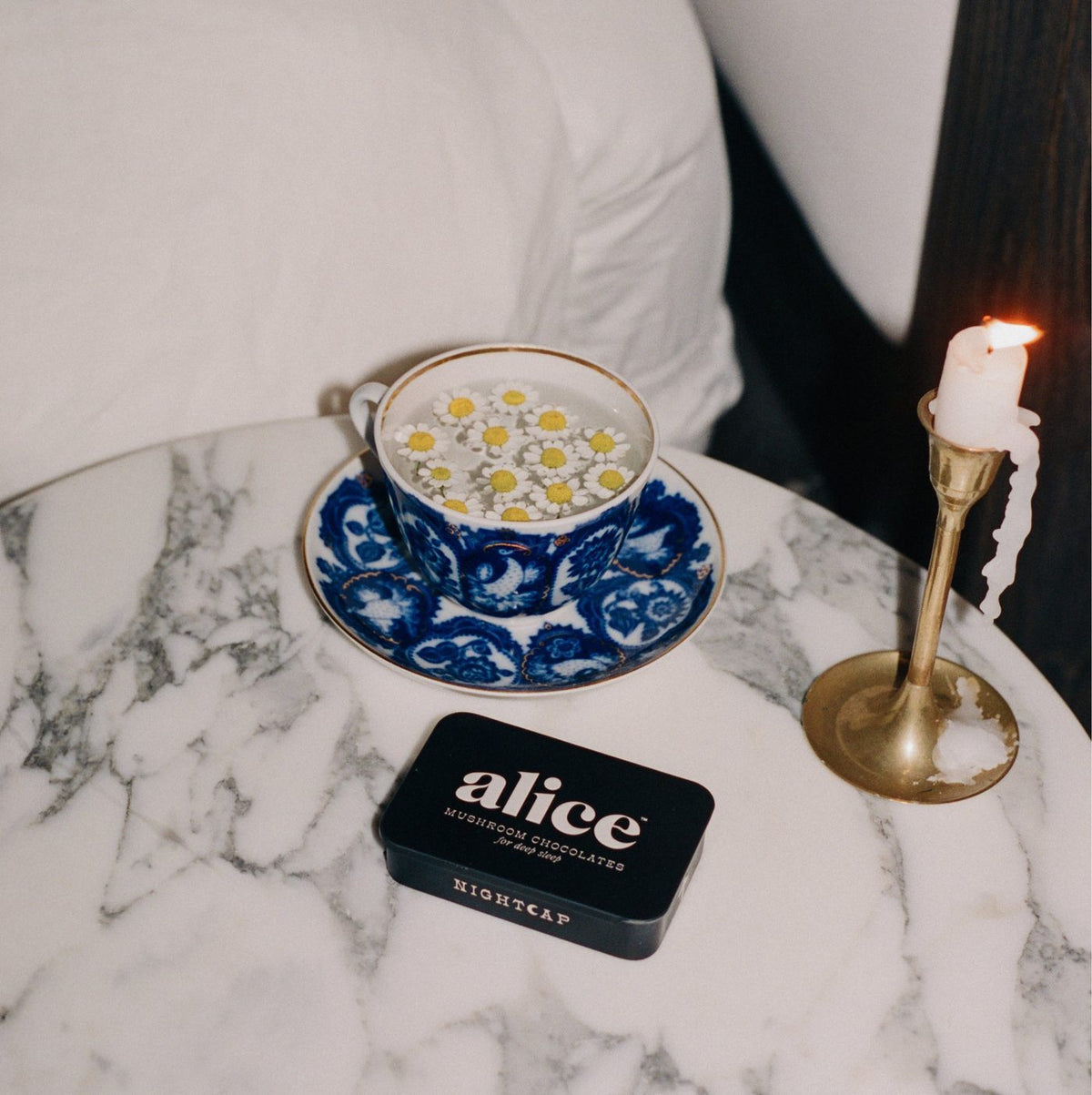 a black nightcap tin on a marble nightstand beside a lit candle, a teacup and a square of dark chocolate